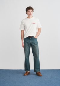 Λευκό κοντομάνικο μπλουζάκι με κόκκιλο λογότυπο «DICKIES», σε συνδυασμό με σκούρα μπλε τζιν παντελόνια φαρδιάς γραμμής και καφέ casual παπούτσια, στέκεται πάνω σε μπλε πάτωμα.