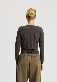 Długi rękaw, ciemnoszara bluzka o gładkiej fakturze, w połączeniu z luźnymi, jasnobrązowymi spodniami z elastycznym paskiem. Widok z tyłu pokazuje dopasowany krój.
