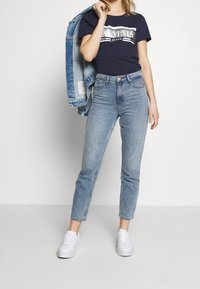 Světle modré džíny s vysokým pasem, tmavě modré tričko Tommy Jeans a bílé tenisky. Džínová bunda je přehozená přes rameno.