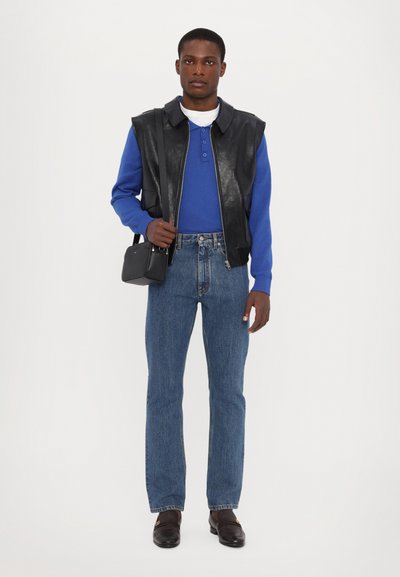 Black leather sleeveless vest over a blue sweater, paired with light blue jeans and dark loafers. Small black handbag accentuates the outfit.