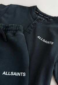 Čierna bavlnená mikina a tepláková súprava s golierom na gombíky a bielym logom "ALLSAINTS" na hrudi a na nohe.