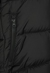 Giacca a vento nera in tessuto, con una tasca con zip. Il materiale è liscio e leggermente lucido, presenta sezioni trapuntate e cuciture a contrasto.