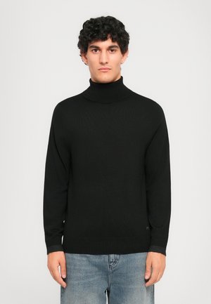 Ung mand med krøllet hår iført en sort turtleneck sweater og lyseblå jeans, stående mod en ensfarvet lys baggrund.
