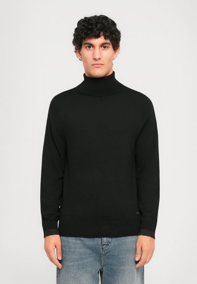 Ung mand med krøllet hår iført en sort turtleneck sweater og lyseblå jeans, stående mod en ensfarvet lys baggrund.