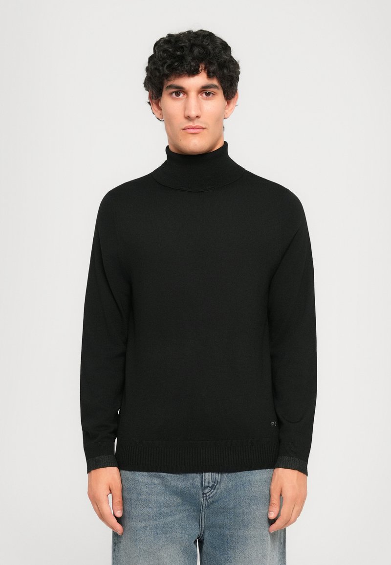 Junger Mann mit lockigem Haar, der einen schwarzen Rollkragenpullover und hellblaue Jeans trägt, steht vor einem schlichten hellen Hintergrund.