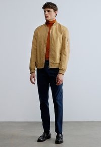 Ljusbeige bomberjacka med ribbade muddar och dragkedja, bärs över en rostfärgad polotröja. Marinblå byxor och svarta skor fullbordar outfiten.