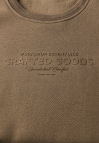 Brun stofskjorte med teksten "WORKSHOP ESSENTIALS CRAFTED GOODS Unmatched Comfort STREET ONE MEN" på brystet.