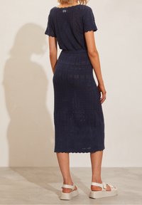 Donna che indossa un vestito midi lavorato a maglia blu navy con maniche corte e sandali con plateau bianchi, in piedi contro un muro beige semplice.