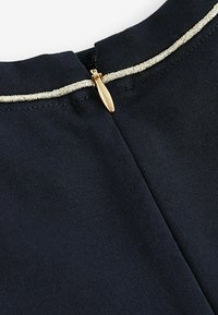 Námořnická modrá látka s texturovaným povrchem, která má zlatý metalický zip a lemování kolem výstřihu pro přidaný detail.