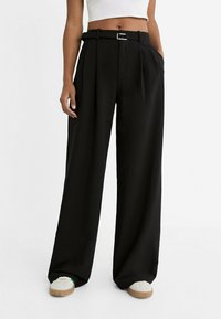 Pantalon noir à jambes larges avec plis, ceinture avec boucle argentée et poches latérales. Porté avec des chaussures blanches et beiges décontractées.