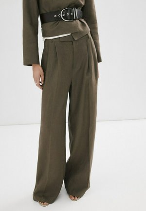 Femme portant un pantalon large olive et une veste assortie avec une large ceinture noire, debout sur un sol blanc.