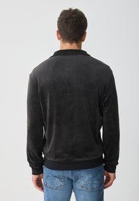 Zwarte, gestructureerde jas met een gladde afwerking, met een geribbelde zoom en manchetten. Draagend over blauwe jeans, achteraanzicht.