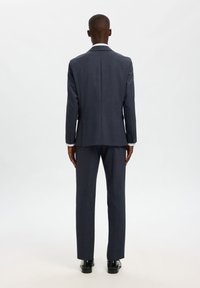 Μπλε σκούρο κοστούμι με υφή, με μονή πίσω σχισμή και παντελόνια στενής γραμμής. Τα παπούτσια είναι μαύρα, με λαμπερό φινίρισμα.