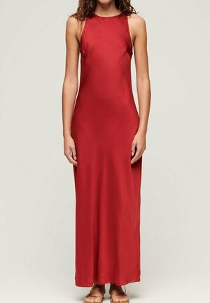 Frau trägt ein ärmelloses, bodenlanges rotes Satinkleid mit hohem Ausschnitt und beigefarbenen Sandalen, steht vor einem schlichten Hintergrund.