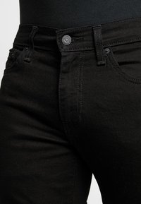 Acercamiento de la cintura de unos jeans de mezclilla negra con botón visible, trabillas para el cinturón y costura del bolsillo delantero.
