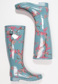 Botas de lluvia florales azules con superficie brillante, con patrones de flores de cerezo rosa y un detalle de hebilla en el lateral. Suela plana para mayor estabilidad.