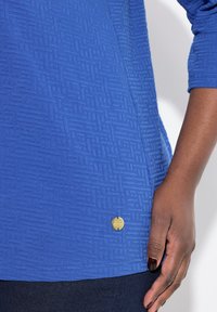 Top azul de manga larga con un patrón geométrico texturizado, con un acento de logo dorado cerca del dobladillo y un acabado de tela suave.