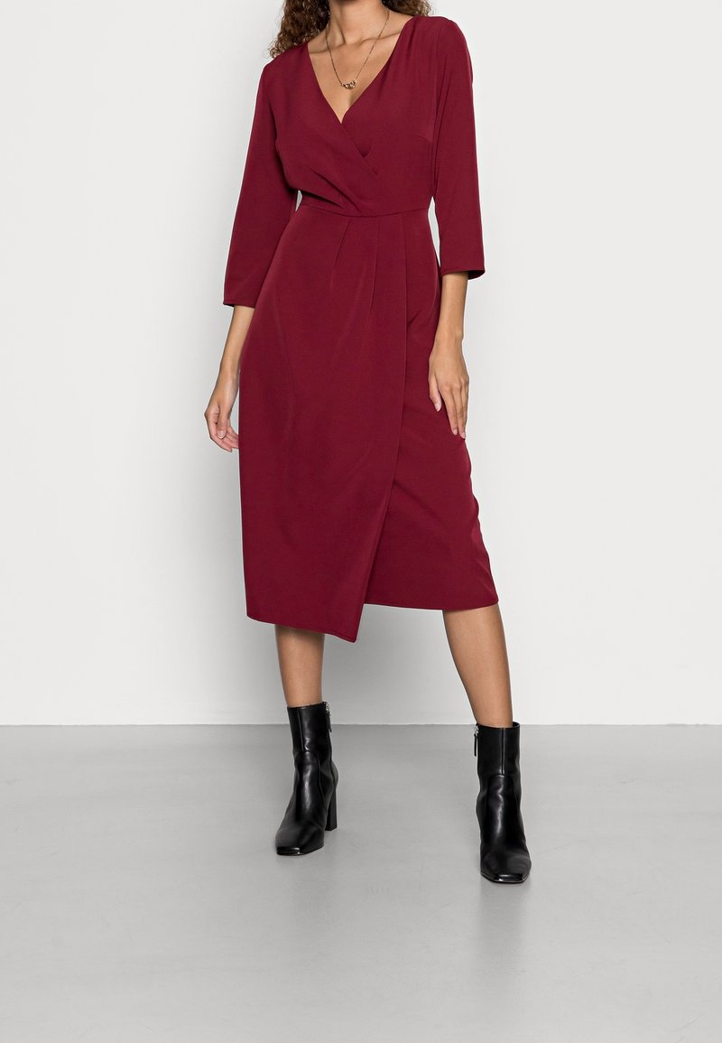 Robe portefeuille bordeaux avec un décolleté en V profond, des manches trois-quarts et une fente à l'avant, associée à des bottines noires à talons. Texture de tissu lisse.