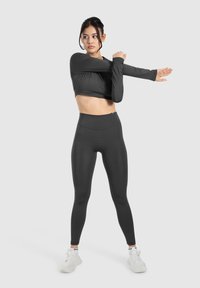 Crop top de sport noir et leggings taille haute, ajustés, texture lisse, mannequin étirant les bras, associé à des baskets blanches.