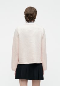 Άτομο με κοντά καστανά μαλλιά που φοράει ένα ανοιχτό ροζ φουσκωτό πουλόβερ πάνω από ένα μπλε καρό πουκάμισο και μια μαύρη πτυχωτή φούστα, γυρισμένο προς την αντίθετη κατεύθυνση της κάμερας.