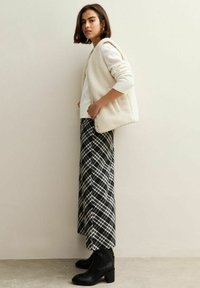 Krämfärgad, fluffig väst över en vit topp, i kombination med svart-vitrutiga byxor och svarta klackboots, stående mot en neutral bakgrund.