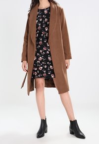 Hnědý dlouhý vlněný kabát se šálovým límcem, nošený přes černé šaty s květinovým vzorem. Modelka kombinovala s černými kotníkovými botami.