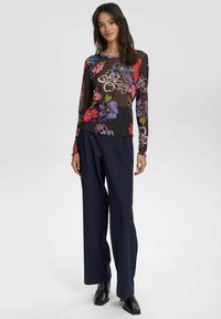 Blusa nera a maniche lunghe con motivi floreali vivaci in rosa, blu e verde; abbinata a pantaloni navy a gamba larga e tacchi neri.