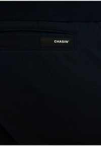 Donkerblauwe stof met een gestructureerde weving, met een zwart label waarop de merknaam "CHASIN'" is gestikt op het achterzakgebied.