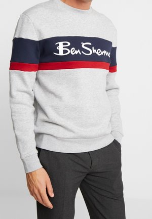 Mężczyzna w szarej bluzie z kapturem z granatowo-czerwonymi poziomymi paskami i logo "Ben Sherman", w połączeniu z ciemnoszarymi spodniami.