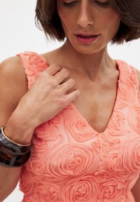 Abito senza maniche color pesca con un motivo floreale testurizzato, caratterizzato da una scollatura a V e rose in tessuto sovrapposte. Accentuato da bracciali scuri sul braccio.