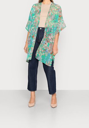 Kvinde iført beige skjorte, mørkeblå jeans, hudfarvede højhælede sko og en løs grøn blomsterprintet kimono-stil cardigan mod en ensfarvet baggrund.