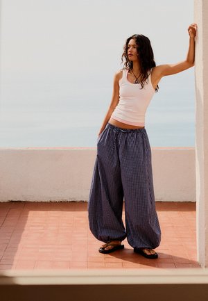 Young woman in loose checkered pants and white tank top stands barefoot, leaning against a white wall on a sunlit terrace by the sea.