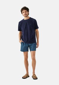 Maglietta in pile a maniche corte blu navy, pantaloni corti turchesi con un motivo testurizzato, indossati con sandali marroni con cinghie. Il modello sta in piedi con le mani nelle tasche.