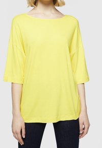 Žluté, oversize, krátké tričko z měkké tkaniny s širokým výstřihem a uvolněným střihem. V kombinaci s tmavými džíny pro kontrast.