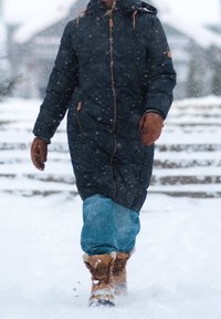 Manteau long et bouffant bleu marine avec une capuche, gants en cuir marron, jean bleu clair et bottes d'hiver beiges, marchant dans la neige pendant une chute de neige.