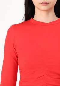 Piros, hosszú ujjú felső, magas nyakkivágással, középen gyűrött textúrával és sima anyaggal, amely formálja a testet.