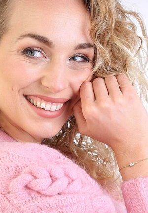 Frau mit lockigem blonden Haar, die einen weichen, rosa gestrickten Pullover trägt. Sie hat ein zartes goldenes Armband mit einem kleinen Edelstein an ihrem Handgelenk.