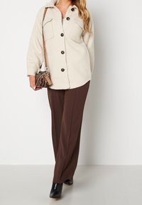 Kurtka polarowa zapinana na guziki w kolorze off-white z dwoma kieszeniami na piersi. Brązowe spodnie z szerokimi nogawkami oraz mała torebka w wzory, zestawione z czarnymi botkami.