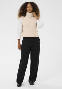 Adīta bezpiedurknē turtleneck džemperis bālā rozā krāsā ar kabelīšu adījuma rakstu un pogu akcentiem, savienots ar melnām platām biksēm un melnām kurpēm.