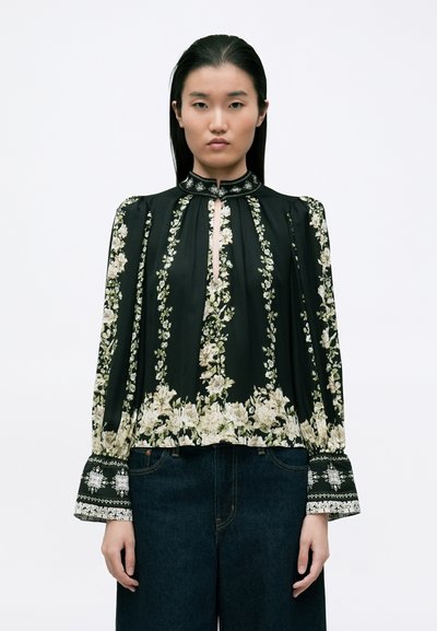 Blouse noire avec des imprimés floraux en crème et vert. Présente un décolleté en forme de clé, des manches bouffantes et des poignets décoratifs avec des motifs complexes.