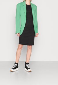 Zielony żakiet na czarnej sukience, połączony z czarnymi sneakersami za kostkę z białymi sznurowadłami i białą podeszwą. Gładki materiał, dopasowany fason.
