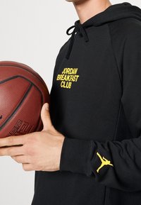 Felpa nera con cappuccio con la scritta "JORDAN BREAKFAST CLUB" in giallo, con un logo Jumpman giallo sulla manica. La persona tiene un pallone da basket.