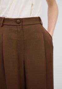 Bruine high-waisted broek met een gladde textuur, voorzien van een sluiting met een voorste knoop, zijzakken en scherpe plooien voor extra detail.