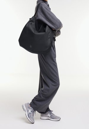 Black soft bag with a rounded shape, made from a smooth material. Includes a logo detail, carried over the shoulder by a person in gray attire.