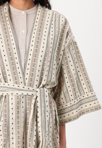 Vzorovaný župan z krémovej látky s zelenými, ružovými a čiernymi pruhmi. S rukávmi v štýle kimono a zodpovedajúcim opaskom. Textúrovaný s vyšívanými akcentmi.