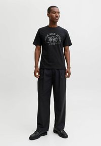 Juodas medvilninis marškinėlis su grafiniu spaudiniu "JACK AND JONES 1990" baltos ir pilkos spalvos. Sujungtas su plačiais juodais kelnėmis ir juodais batais.