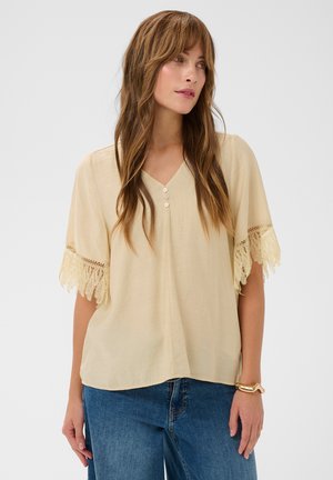 Moteris su ilgais rudais plaukais, dėvinti smėlio spalvos palaidinę su nėrinių apdaila ant rankovių ir mėlynas džinsas, stovinti priešais paprastą foną.