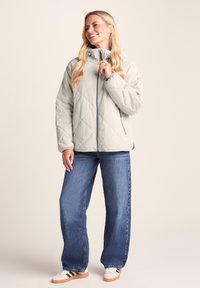 Light gray quilted jacket with high collar, zip closure, and side pockets, paired with loose blue jeans and white sneakers.