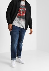 Cardigan nero con zip e finitura texturizzata, t-shirt grigia con grafiche e dettagli rossi, jeans blu e sneakers bianche con risvolti.