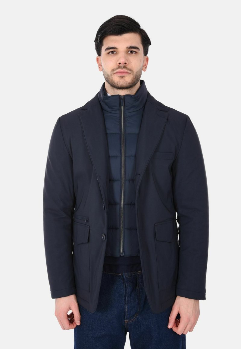 Giacca due-in-uno blu navy con interno trapuntato testurizzato, doppio bottone frontale e due tasche anteriori. Strato esterno liscio e sartoriale.